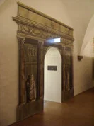 Das Bild zeigt eine Tür, die in eine weiße Wand eingelassen ist und einen eleganten und künstlerischen Eingang zu dem schafft, was wie ein Ausstellungs- oder Museumsraum aussieht. Die Türöffnung wird von einem gedruckten Design eingerahmt, das das Aussehen der klassischen Architektur nachahmt.