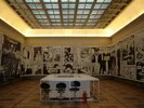 Das Bild zeigt einen weitläufigen Raum, der nach der Art der Ausstellung und der Beleuchtung wie ein Museum oder eine Galerie aussieht. Der Raum zeichnet sich durch eine lange, geschwungene Wand aus, die mit einer großformatigen monochromen Installation geschmückt ist. Dieses Kunstwerk setzt sich aus zahlreichen Fotografien zusammen, die in Tafeln angeordnet sind.