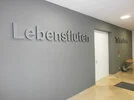 An der Wand sind zwei Schilder angebracht: "Lebensfluten" und "Tatensturm". Ein weißes Türblatt ist ebenfalls zu sehen. Der Gesamteindruck ist sachlich und minimalistisch.