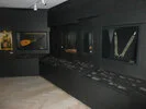 Das Bild zeigt einen Ausstellungsraum mit dunklen Wänden und einem hellen Holzfußboden. In Vitrinen sind verschiedene Exponate ausgestellt, darunter eine Laute, ein Paar Hosenträger und diverse kleinere Gegenstände. Die Vitrinen sind mit Beschriftungen versehen. Der Raum wirkt modern und minimalistisch gestaltet.
