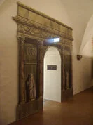 Das Bild zeigt eine Tür, die in eine weiße Wand eingelassen ist und einen eleganten und künstlerischen Eingang zu dem schafft, was wie ein Ausstellungs- oder Museumsraum aussieht. Die Türöffnung wird von einem gedruckten Design eingerahmt, das das Aussehen der klassischen Architektur nachahmt.