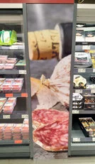  Im Vordergrund steht ein großes Werbeposter mit Bildern von Salami, das zwischen zwei Kühlregalen platziert ist. 
