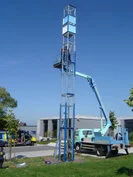 Arbeiter montieren eine hohe Metallstruktur mit Hilfe eines blauen Hebebühnenfahrzeugs auf einem Firmengelände